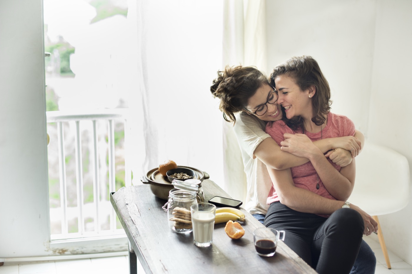 Two women embracing and laughing at a sunny kitchen table